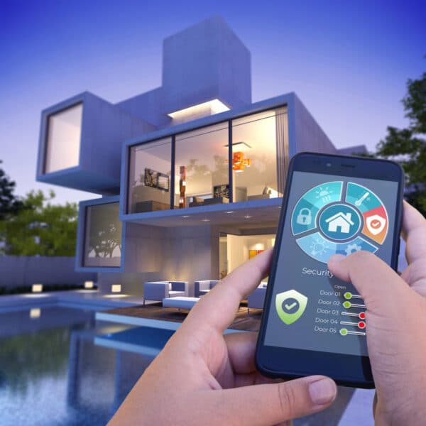 A hand holding a smartphone with smart home controls in front of a modern smart residential home at twilight
