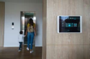 A tablet showing smart home controls mounted on a wood-paneled wall with a parent and kid in the background