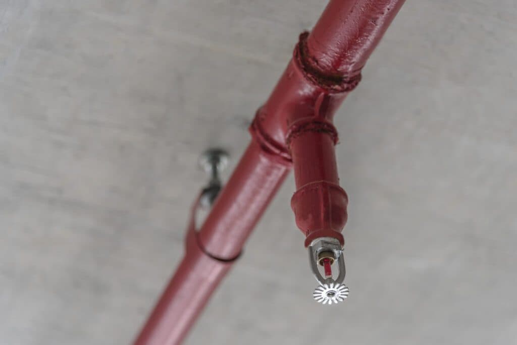 A red fire sprinkler head mounted under a grey concrete ceiling next to a red pipe