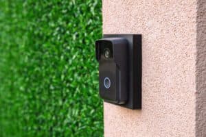 A smart video doorbell unit mounted on a textured pink stucco wall next to artificial grass paneling