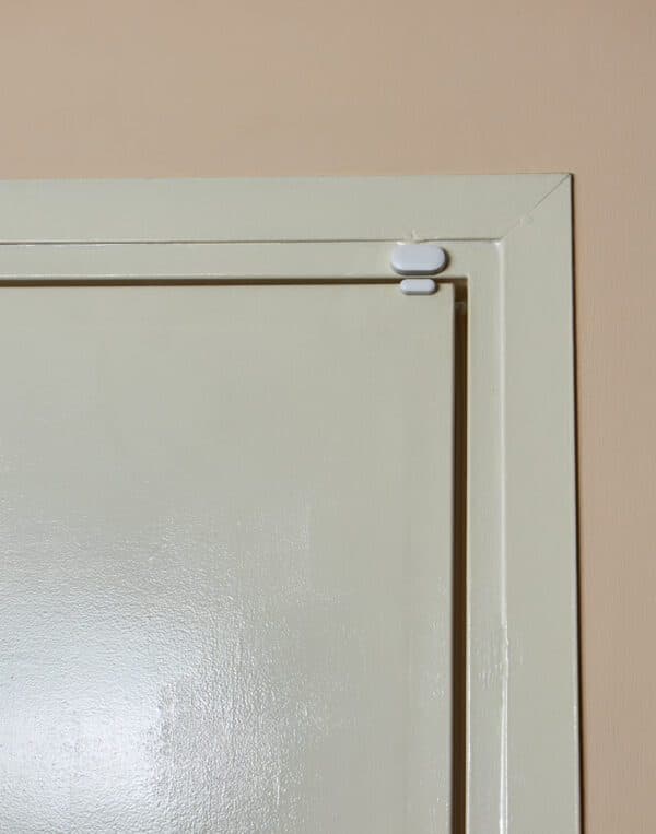 A door sensor transmitter mounted on the top corner of a closed cream-colored interior door frame