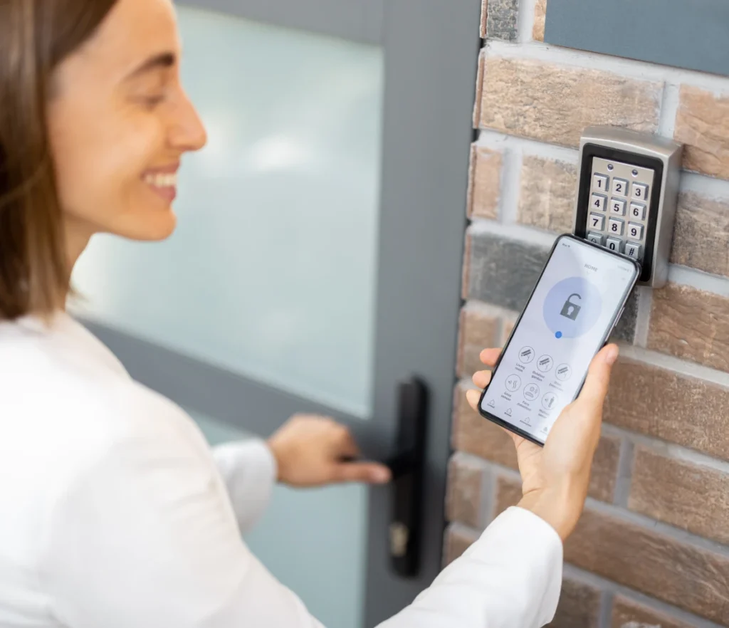 A smiling woman using her smartphone to unlock a smart wall-mounted keypad next to a door