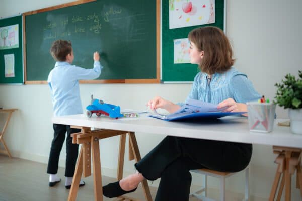 Teacher working at desk with elementary school student writing on blackboard